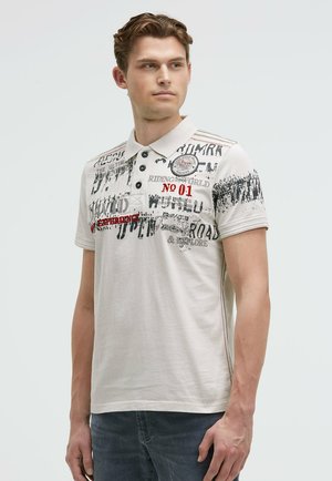 Hellcreme Polo-Shirt mit schwarzen und roten Drucktexten, dreiknöpfiger Knopfleiste, kurzen Ärmeln und einer Seitennaht mit roten Stichdetails.