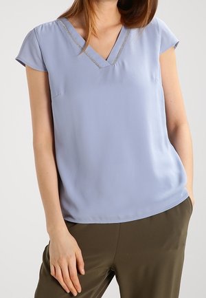 Mujer con blusa azul claro de manga corta con escote en V y ribete plateado y pantalones verde oliva, de pie con una mano en el bolsillo.