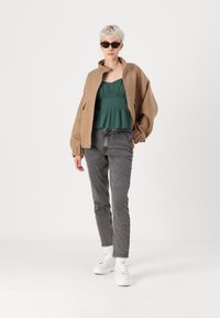 Hnědá oversized bunda, zelený volný top, šedé džíny a bílé tenisky. Doplňky zahrnují černé sluneční brýle a kruhové náušnice.