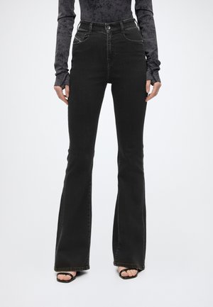 Persona che indossa jeans neri a vita alta e gamba svasata con tasche frontali, abbinati a sandali neri aperti e un top scuro a maniche lunghe con fantasia.