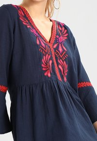 Blusa blu navy con scollo a V, caratterizzata da vivaci ricami floreali rosa e rossi lungo il collo e le maniche, e tessuto testurizzato.