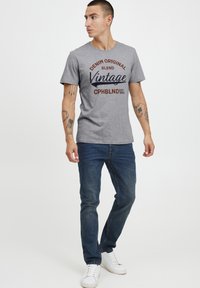 Šedé bavlněné tričko s potiskem "DENIM ORIGINAL BLEND Vintage CPHBLND EST. 1993" v různých barvách. V kombinaci s modrými džínami a bílými teniskami.
