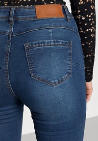 Close-up de uma pessoa a usar jeans de denim azul escuro com um remendo de couro rotulado "NOISY MAY" na cinturinha e uma blusa de renda preta.