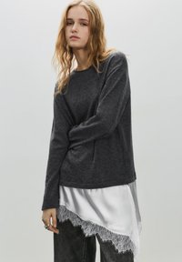 Top tricotat cu mânecă lungă în culoare gri închis peste o bluză albă din satin cu margine de dantelă. Modelul poartă blugi închiși la culoare; fundal neutru.