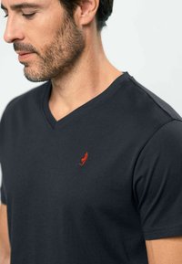 Juoda V formos kaklo marškinėliai iš minkštos medžiagos, su mažyčiu raudonu siuvinėjimu kairėje krūtinės pusėje. Trumpomis rankovėmis ir prigludusio silueto.