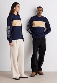 Jūras spēku melnas kreklu adījuma sporta jakas ar bēšām horizontālām svītrām, ar "LACOSTE" logo uz piedurknēm. Saskaņotas ar gaismu un tumšu platā kāju biksēm.