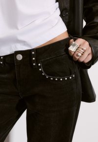 Čierne denimové džínsy so striebornými ozdobnými cvočkami na vreckách a páse. Nosí sa s bielym tričkom a striebornými prsteňmi. Detailný pohľad na oblečenie.
