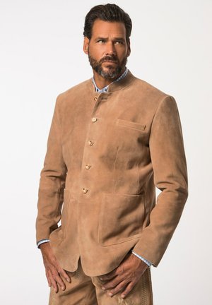Chaqueta de ante color arena con cuello alzado, que presenta cuatro botones frontales, dos bolsillos de parche y costuras sutiles. Llevada sobre una camisa a cuadros azules.