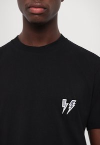 Mand iført en ensfarvet sort t-shirt med to hvide lynsymboler på venstre bryst mod en ensfarvet baggrund.