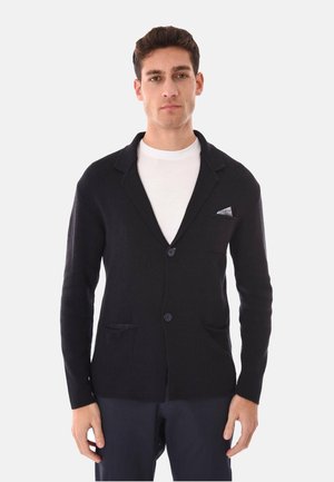 Uomo che indossa un cardigan nero abbottonato sopra una camicia bianca, in piedi davanti a uno sfondo chiaro uniforme, rivolto verso l'obiettivo con espressione neutra.