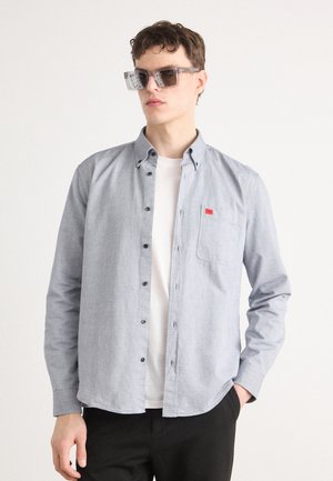 Lichtblauw button-down shirt van een stof met een zachte textuur, met een borstzak en een klein rood label. Gedragen over een wit t-shirt.