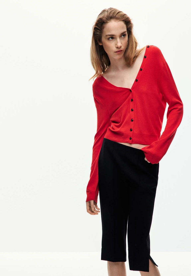 Jeune femme portant un haut rouge ample boutonné, tombant sur une épaule, et un pantalon noir court, debout devant un fond clair uni.