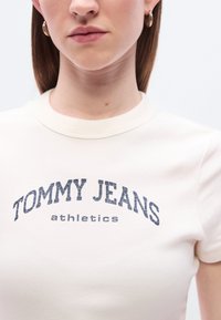 Κρεμ χρωματιστό T-shirt με εφαρμοστή σχεδίαση, που διαθέτει γραφικό κείμενο σε σκούρο μπλε χρώμα "TOMMY JEANS athletics" με μια ελαφρώς φθαρμένη γραμματοσειρά.