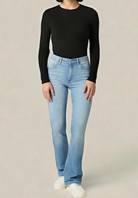 Femme portant un haut noir à manches longues, un jean taille haute bleu clair et des baskets blanches, debout devant un fond neutre uni.