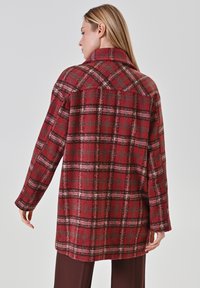 Donna che indossa un lungo cappotto a quadri rosso, rosa e grigio, con le braccia rilassate, di spalle, su uno sfondo grigio chiaro uniforme.