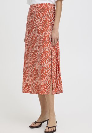 Naine, kes kannab põlveni ulatuva oranži-beeži mustriga seelikut, millel on külje lõhik, pruunide kontsaga sandaalide ja valge topiga.