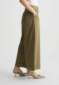 Pantaloni a gamba larga verde oliva con tasche laterali, taglio sartoriale e orli risvoltati. Il materiale appare leggero e liscio al tatto.