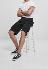 Černé cargo šortky s několika kapsami, délka ke kolenům a nastavitelnými bočními stahovacími šňůrkami; spojené s černými teniskami a bílým tričkem.