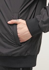 Black bomber jacket with smooth texture, elastic cuffs, and side pocket, showcasing a hand resting casually inside the pocket.