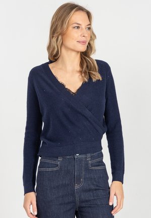 Kobieta w granatowym swetrze z dzianiną w kształcie płaszcza z koronkowym wykończeniem jako warstwą wewnętrzną oraz ciemnych, wysokich dżinsach, patrząca na bok na tle jednolitego tła.