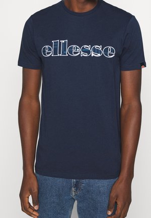 Mężczyzna w granatowej koszulce z białym i niebieskim napisem "ellesse" na piersi oraz w niebieskich dżinsach, stojący na jednolitym tle.