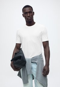 Άνδρας με λευκό μπλουζάκι και γκρι πουλόβερ δεμένο γύρω από τη μέση, κρατώντας μαύρο καπέλο στο χέρι, στέκεται μπροστά σε απλό ανοιχτόχρωμο φόντο.