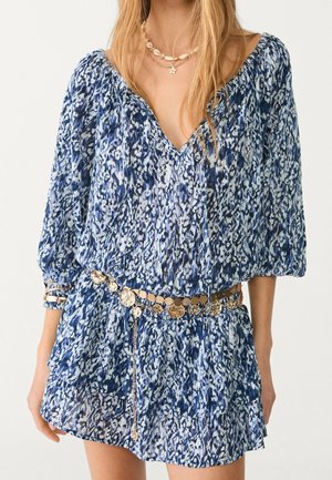 Blue and white leopard print dress with long puff sleeves, deep V-neck, gold coin belt, and layered gold necklaces on a woman.