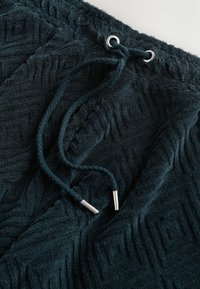 Tejido texturizado de color azul oscuro con un patrón geométrico. Presenta un cordón con extremos de metal y una superficie suave y mullida.