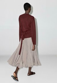 Maroon sweater with a tie at the waist, paired with a light gray pleated skirt. The model wears black sandals. The background is neutral.