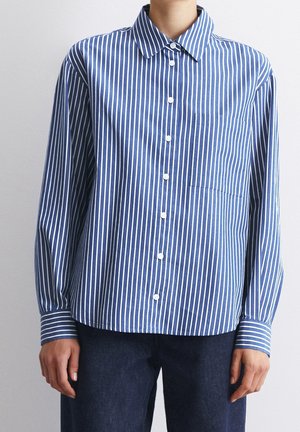 Persoon draagt een blauw shirt met lange mouwen met witte verticale strepen, knopen aan de voorkant en een enkele borstzak, gecombineerd met een donkere broek.