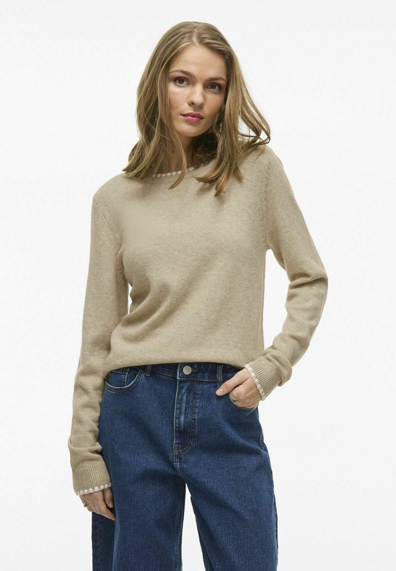 Suéter beige de punto con mangas largas, puños acanalados y cuello redondo, combinado con jeans de mezclilla azul de talle alto.