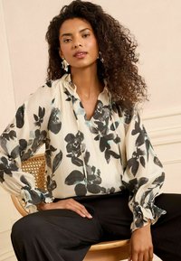 Floral blouse in ivory with black and blue flower patterns, puffy sleeves with elastic cuffs, and a loose fit. Paired with black pants.