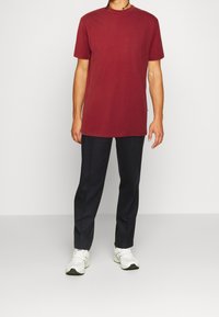 Bordeauxfarbiges Baumwoll-T-Shirt mit Rundhalsausschnitt, kombiniert mit navyblauen Hosen und weißen Sportschuhen. Verfügt über eine lockere Passform und kurze Ärmel.