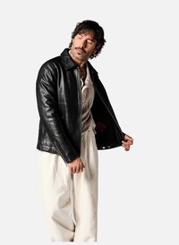 Veste en cuir noir avec un design à col, fermeture éclair sur le devant et poches latérales. Portée par-dessus un haut à motifs de couleur claire et un pantalon blanc.