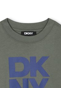 Olijfgroene katoenen t-shirt met een ronde hals en een groot blauw "DKNY" logo op de voorkant. De stof lijkt zacht met een gladde textuur.