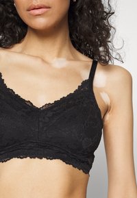 Čierna čipkovaná bralette s výstrihom do V, zdobená kvetinovými motívmi, jemnými zúbkovanými okrajmi a tenkými nastaviteľnými ramienkami. Mäkká textúrovaná látka.