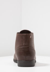 Botas de cuero marrón hasta el tobillo con una textura suave, punta redonda y tacón bajo de goma negra. Presentan costuras y un pequeño logotipo en la parte trasera.