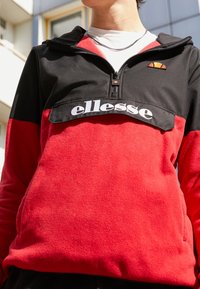 Sudadera de forro polar en negro y rojo, con media cremallera, logo bordado y acabado texturizado. Incluye un pequeño detalle en naranja sobre el logo.