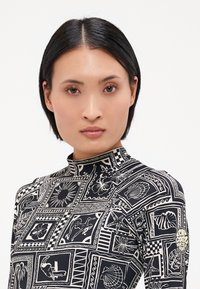 Haut à manches longues noir et blanc avec un col haut, présentant un motif géométrique de coquillages et de fleurs. Comprend un petit logo sur la manche.