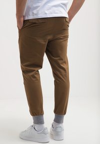 Pantaloni marroni a gamba affusolata in tessuto liscio, con polsino elasticizzato, tasche laterali e vestibilità casual, abbinati a sneakers bianche.