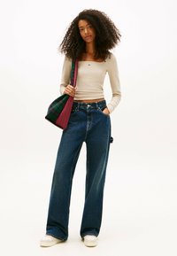 Mujer con cabello rizado que lleva una camiseta de manga larga beige corta, jeans de pierna ancha azules, zapatillas blancas y carga una bolsa de tote de cuadros verdes y rojos.