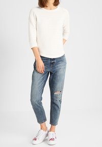 Haut blanc texturé à manches longues, jeans bleus déchirés, et baskets blanches avec accents floraux, debout sur un fond neutre.