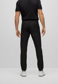 Čierne elastické nohavice so slim fit strihom, ktorý má dve zadné vrecká a hladkú textúru. Model má na sebe biele tenisky pre kontrast.