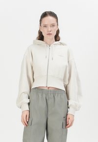 Korte beige hoodie met een ritssluiting, grote pofmouwen en een klein logopatch op de borst, gecombineerd met losse grijze cargobroeken.