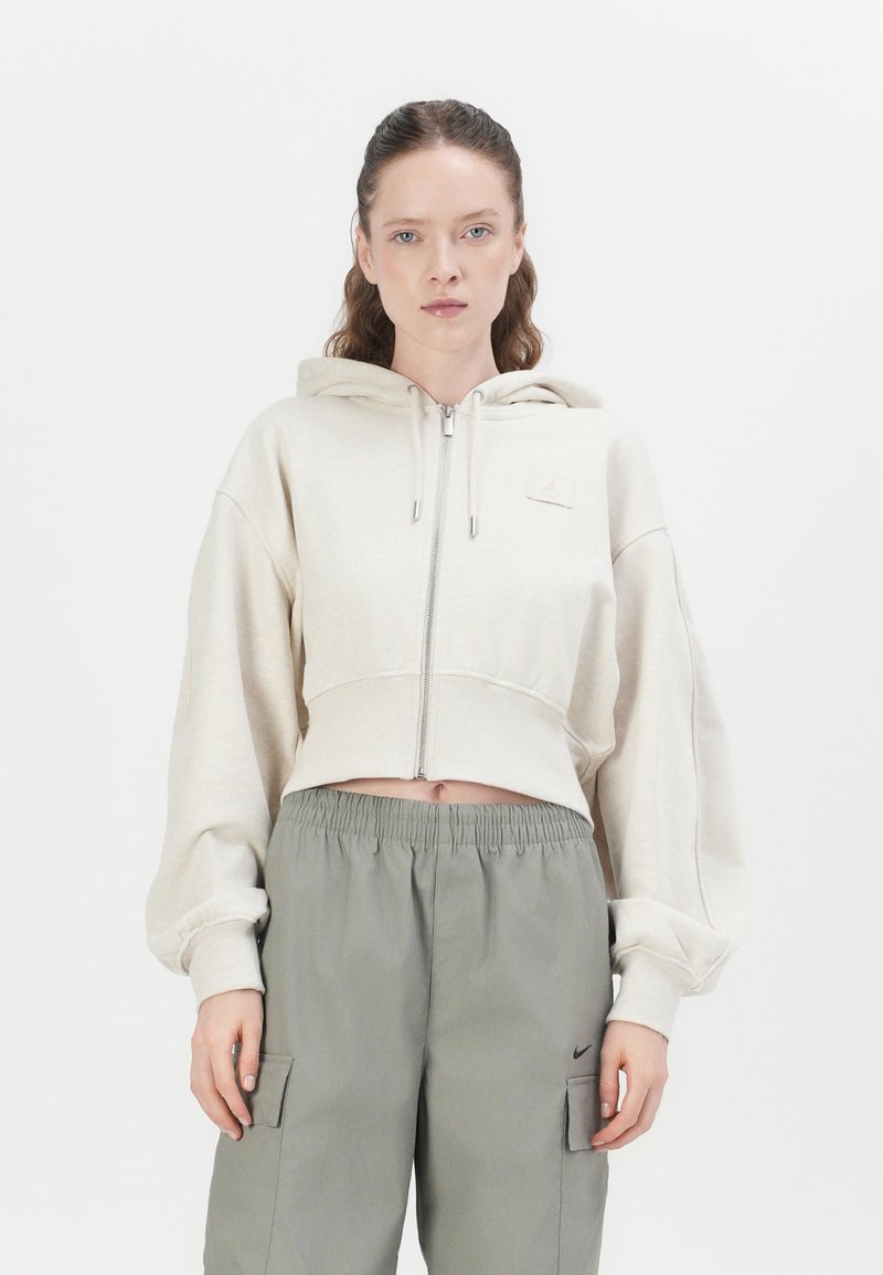 Korte beige hoodie met een ritssluiting, grote pofmouwen en een klein logopatch op de borst, gecombineerd met losse grijze cargobroeken.