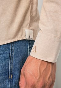 Beżowa koszula z subtelną teksturą, z metką oznaczoną „JP AWARE” oraz wyszywanym trójkątnym detalem na mankiecie. Noszona z niebieskimi dżinsami.