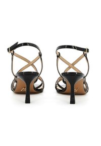 Tacchi in vernice nera con un design a fascia incrociata, sottile cinturino alla caviglia e tacco a stiletto di 7 cm. Le solette color nude contrastano con l'esterno scuro.