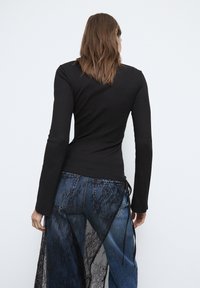 Zwarte top met lange mouwen gecombineerd met blauwe spijkerbroek, voorzien van een doorschijnende zwarte kanten overlay. De spijkerbroek heeft gestrikte accenten aan de achterkant.