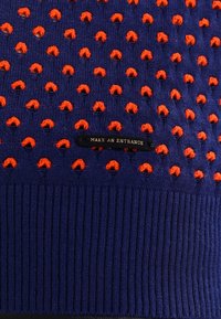 Pletený svetr v tmavě modré barvě s vyvýšenými oranžovými bodovými vzory; má ribbovaný lem a štítek s nápisem "UDĚLEJTE VSTUP."