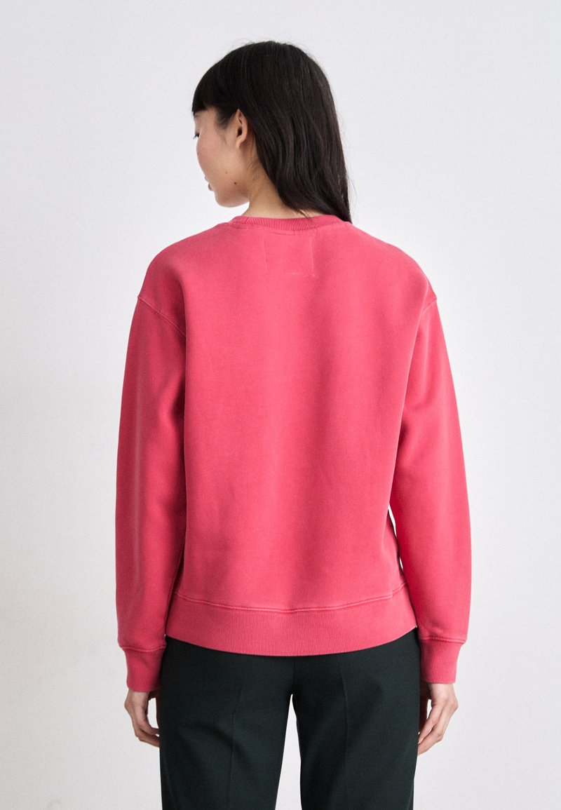 Czerwony sweatshirt z okrągłym dekoltem o luźnym kroju, z prążkowanymi mankietami i dołem. Miękka tekstura materiału. Noszony z ciemnymi spodniami.